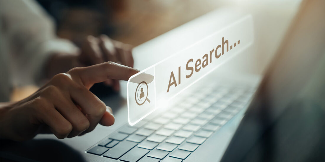 Realistic image of a person’s hand resting on a laptop keyboard with one finger of another hand approaching a search bar with the words “AI Search.”