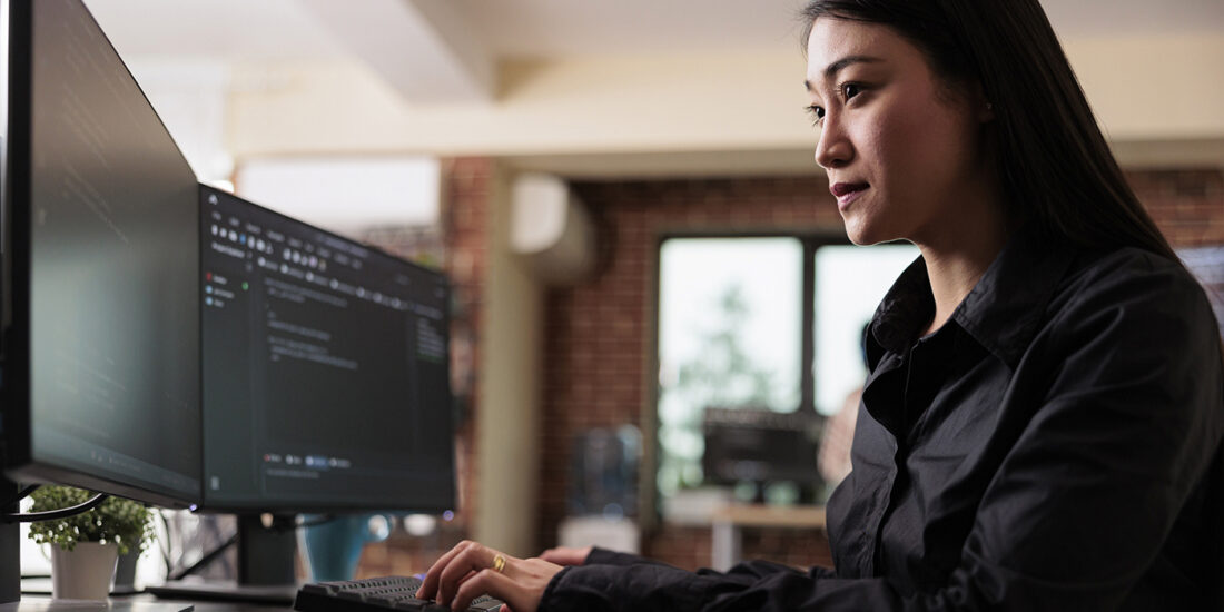 The image shows an Asian woman focused on her work at a modern, multi-monitor computer workstation. She is wearing a dark blouse, and her workspace is well-lit with natural light, suggesting a comfortable and contemporary office environment. Her posture and the concentration on her face indicate she is deeply engaged in a task, possibly related to cybersecurity, given the content on the screens.