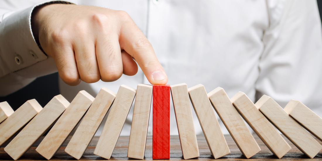 The image shows a close-up of a person's hand pushing a red domino, which stands out among a sequence of white dominoes set on a wooden surface. The action captures the moment before the red domino is likely to fall and initiate a chain reaction with the other dominoes. The person is wearing a white shirt, and only their hand is visible. The background is neutral and out of focus, emphasizing the dominoes and the action.