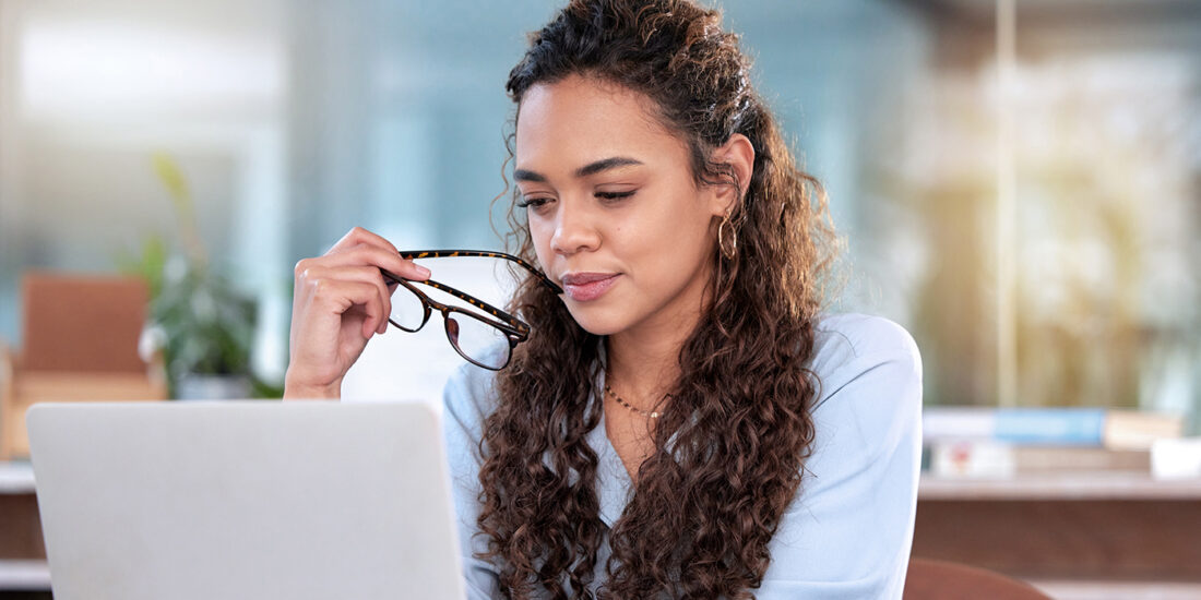 The image depicts a woman sitting in front of a laptop. She appears focused and contemplative, holding a pair of glasses in her hand while looking at the screen. Her hair is styled in long, curly locks that cascade down her shoulders. She wears a light blue blouse, which suggests a professional or business casual environment. The setting appears to be a modern, well-lit office with a blurred background, indicating a possible workspace.
