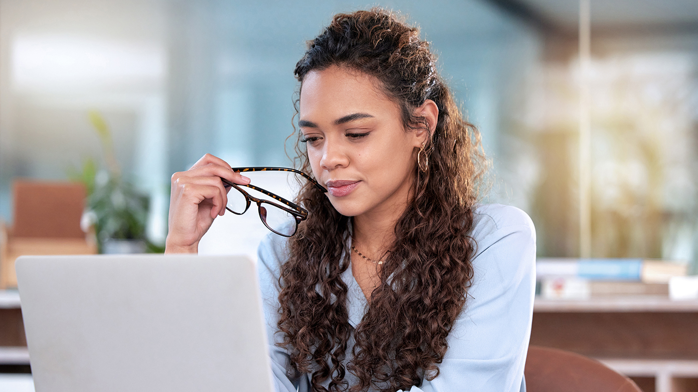 The image depicts a woman sitting in front of a laptop. She appears focused and contemplative, holding a pair of glasses in her hand while looking at the screen. Her hair is styled in long, curly locks that cascade down her shoulders. She wears a light blue blouse, which suggests a professional or business casual environment. The setting appears to be a modern, well-lit office with a blurred background, indicating a possible workspace.