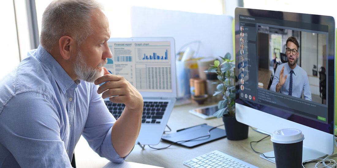 A mature businessman with a thoughtful expression is participating in a video conference on his desktop computer. The screen shows a younger man gesturing as if explaining something. The businessman's desk features an open laptop displaying financial charts, a notepad, and a closed cup of coffee, indicating a professional setting. This image reflects how businesses might utilize virtual Chief Information Officer (vCIO) services to receive remote, strategic consultation on IT matters, exemplifying why to use a vCIO for expertise without the need for an in-house executive.