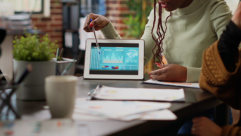 Two professionals are engaged in strategic planning at a casual office setting, focusing on a tablet displaying colorful data charts and graphs. One person, who appears to be explaining the data, could represent a Virtual Chief Information Officer (vCIO) providing expert analysis and advice on technology strategy. This scenario exemplifies virtual chief information officer services, illustrating the benefits of why to use a vCIO for businesses seeking flexible, high-level IT leadership and insights.