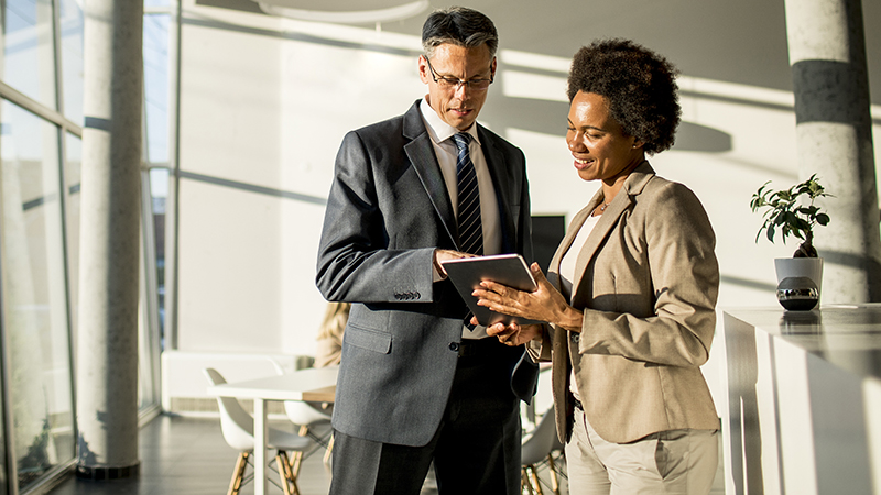 In a sunlit modern office with floor-to-ceiling windows, a well-dressed man and woman are in mid-discussion, with the man attentively looking at a tablet the woman is holding. This could be a depiction of a business consultation, where the woman, possibly embodying a Virtual Chief Information Officer (vCIO), is providing specialized virtual chief information officer services. They are collaborating to enhance the company's technological strategy, showing why a vCIO is essential for forward-thinking IT management.