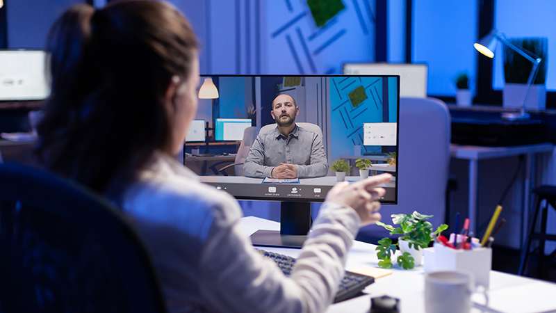 A woman is at her desk in a modern office environment during nighttime, engaged in a video call with a man on her computer screen. The man appears to be speaking with confidence and could represent a Virtual Chief Information Officer (vCIO) providing remote guidance and virtual chief information officer services. This image illustrates the convenience and effectiveness of a vCIO in action, emphasizing the benefits of why to use a vCIO for strategic IT decision-making and support.