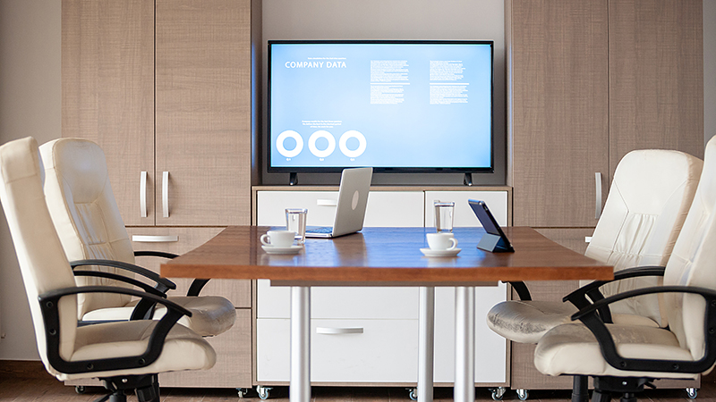 A contemporary conference room showcasing technology trends with a large display screen presenting company data. The room features a rectangular wooden table with ergonomic office chairs around it, a laptop and a tablet set up for a presentation, and coffee cups ready for participants. The room's design is minimalistic with a neutral color palette, blending functionality with modern aesthetics.