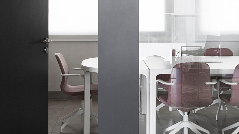 sleek, modern conference room viewed through a partially opened door, highlighting technology trends with clean design lines. The room is furnished with stylish pink and white office chairs positioned around a minimalist white table, possibly equipped with connectivity ports for digital devices. The glass walls of the room suggest an open and collaborative space, while the uncluttered setup promotes a focused and high-tech meeting environment.