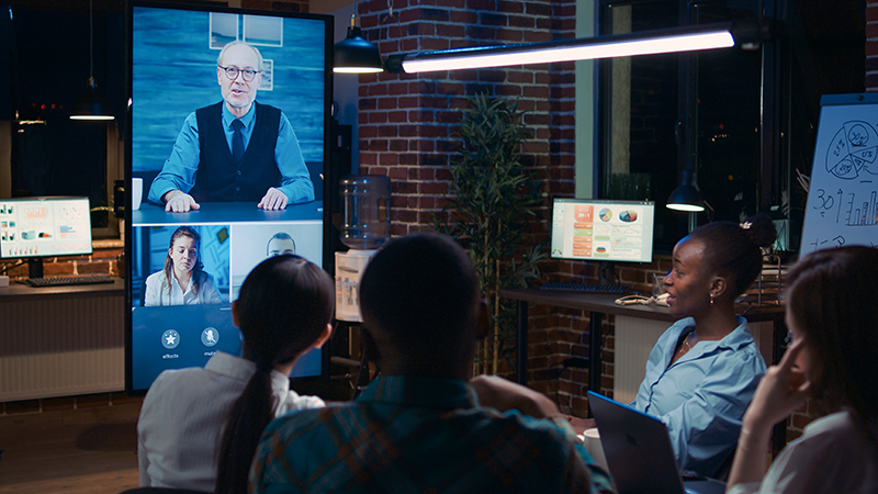 A group of professionals in a trendy, brick-walled conference room engaging with cutting-edge technology trends during a virtual meeting. The central focus is a large digital screen where a man in business attire is speaking, supported by smaller screens showing additional participants. The room is casually furnished, blending the warmth of an exposed brick ambiance with the modernity of advanced communication tools. Overhead lighting and a relaxed evening atmosphere add to the innovative and collaborative workspace.