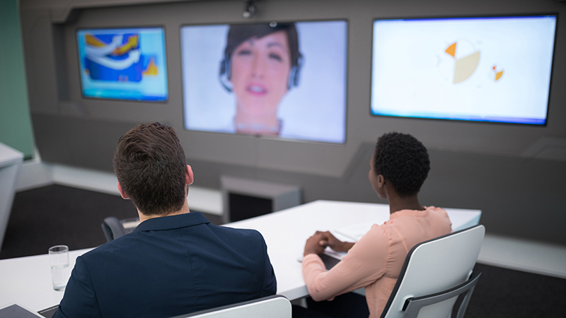 Two professionals seated at a sleek white conference table in a modern room, focused on presentations displayed across three large screens as part of the latest conference room technology trends. Each screen shows different content, including graphical data and a live video feed of a female presenter, demonstrating the capacity for dynamic and interactive virtual meetings. The setting exemplifies a high-tech, efficient workspace designed for collaborative digital engagement.
