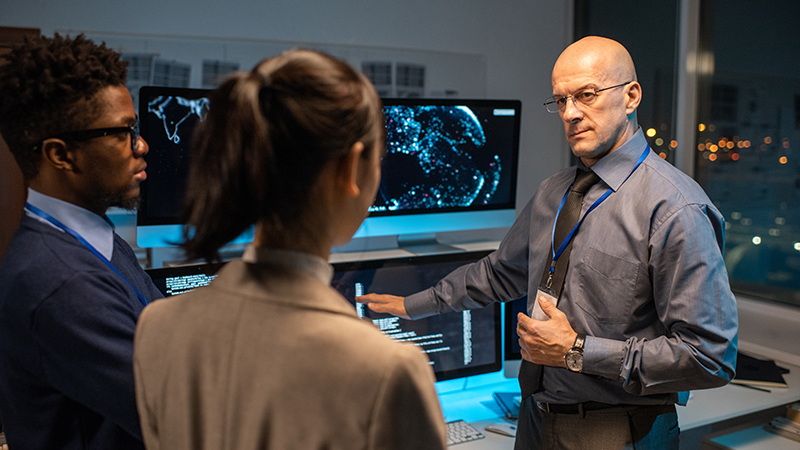 Three cybersecurity professionals are analyzing the cybersecurity landscape on multiple computer screens in a dimly lit control room. The team, consisting of two men and one woman, are focused on a map display, with one man pointing to a screen, possibly identifying a threat or area of interest. They are all wearing business attire, indicating an executive environment. 