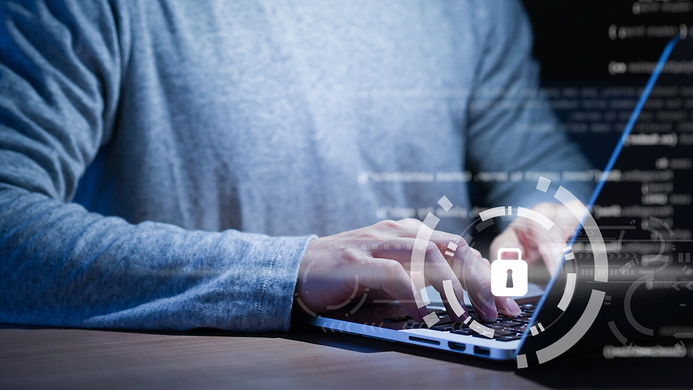 A person in a gray long-sleeved shirt is typing on a laptop with digital graphics of a lock symbol overlaid on the image, symbolizing vigilance in the cybersecurity landscape.