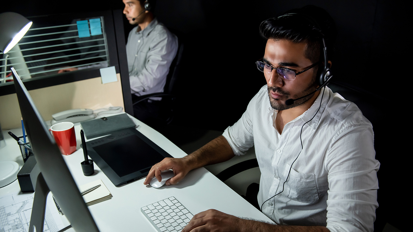 A man wearing a headset with a microphone sits at a managed IT help desk in a dimly lit office environment. He is focused on a computer monitor, operating a mouse and keyboard, with a digital drawing tablet, a red cup, and office supplies on the desk. Another person with a headset can be seen in the background, suggesting a collaborative workplace.