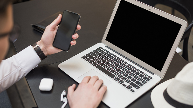 A person at a work desk holding a smartphone in one hand while the other hand is on a laptop's touchpad, with a pair of earbuds and a charging case beside the laptop. The smartphone and laptop potentially represent shadow IT risks in a 'bring your own device' work environment.