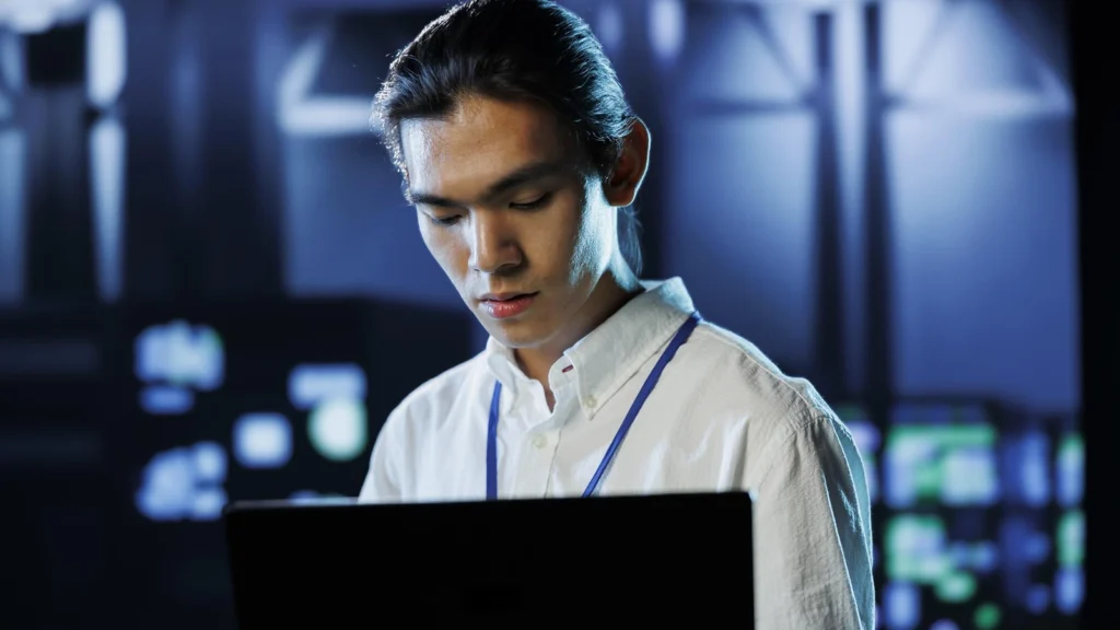A young IT professional wearing a lanyard, intently working on a laptop in a high-tech environment, with blurred server lights in the background. A young IT professional wearing a lanyard, intently working on a laptop in a high-tech environment, with blurred server lights in the background.