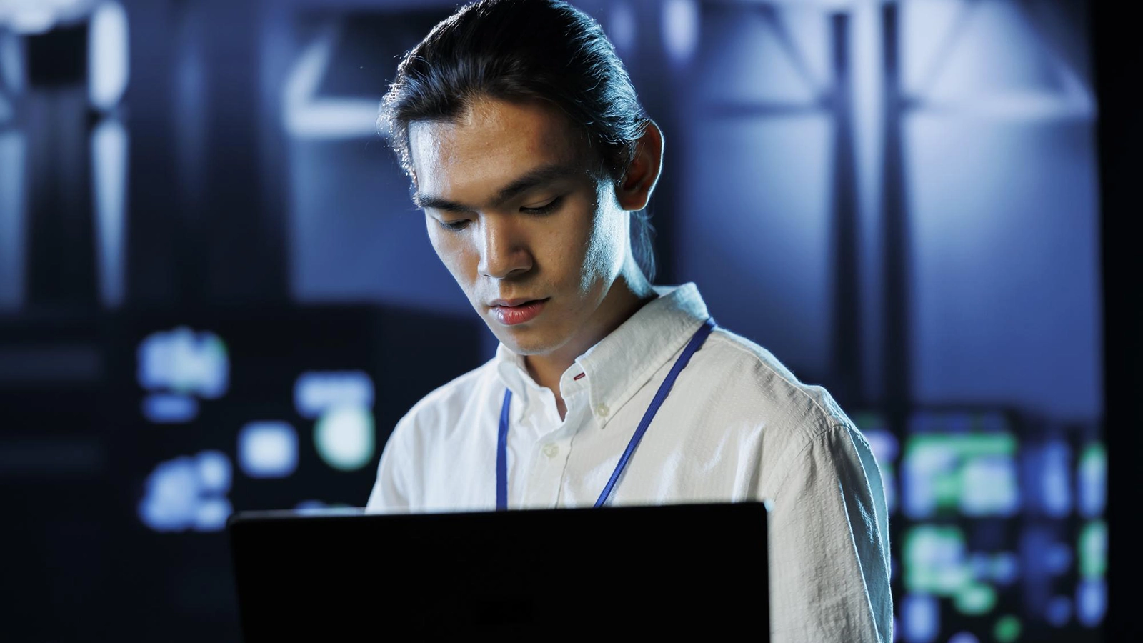 A young IT professional wearing a lanyard, intently working on a laptop in a high-tech environment, with blurred server lights in the background.