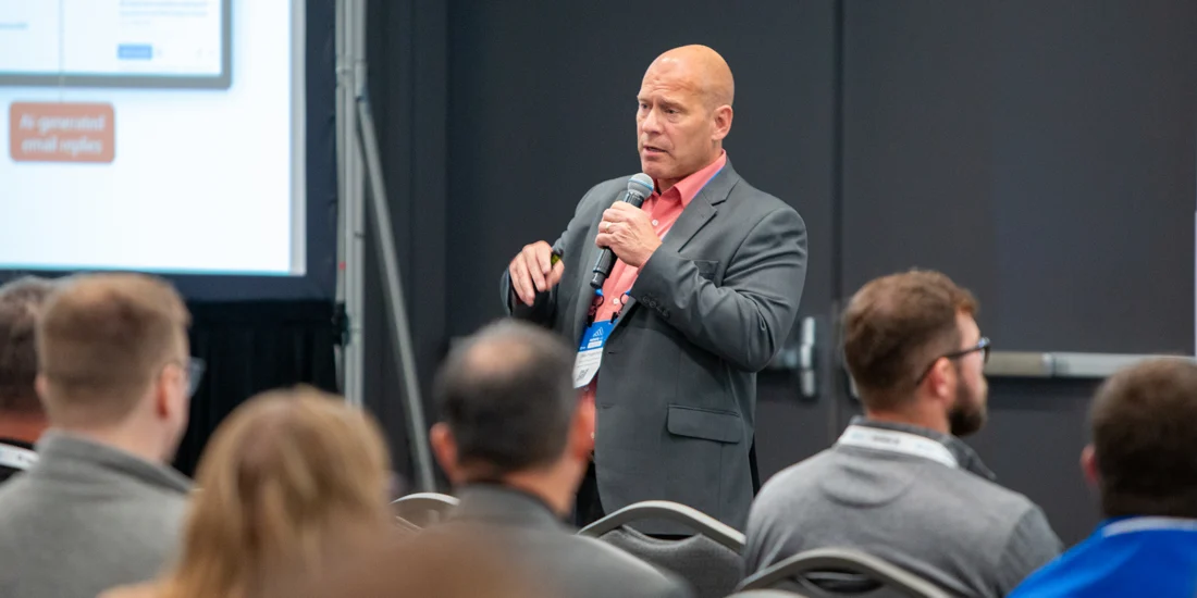 A speaker presenting at the Microsoft Power Platform Envisioning Workshop. The presenter, wearing a suit and holding a microphone, addresses an audience seated in rows while a screen with slides is partially visible in the background.
