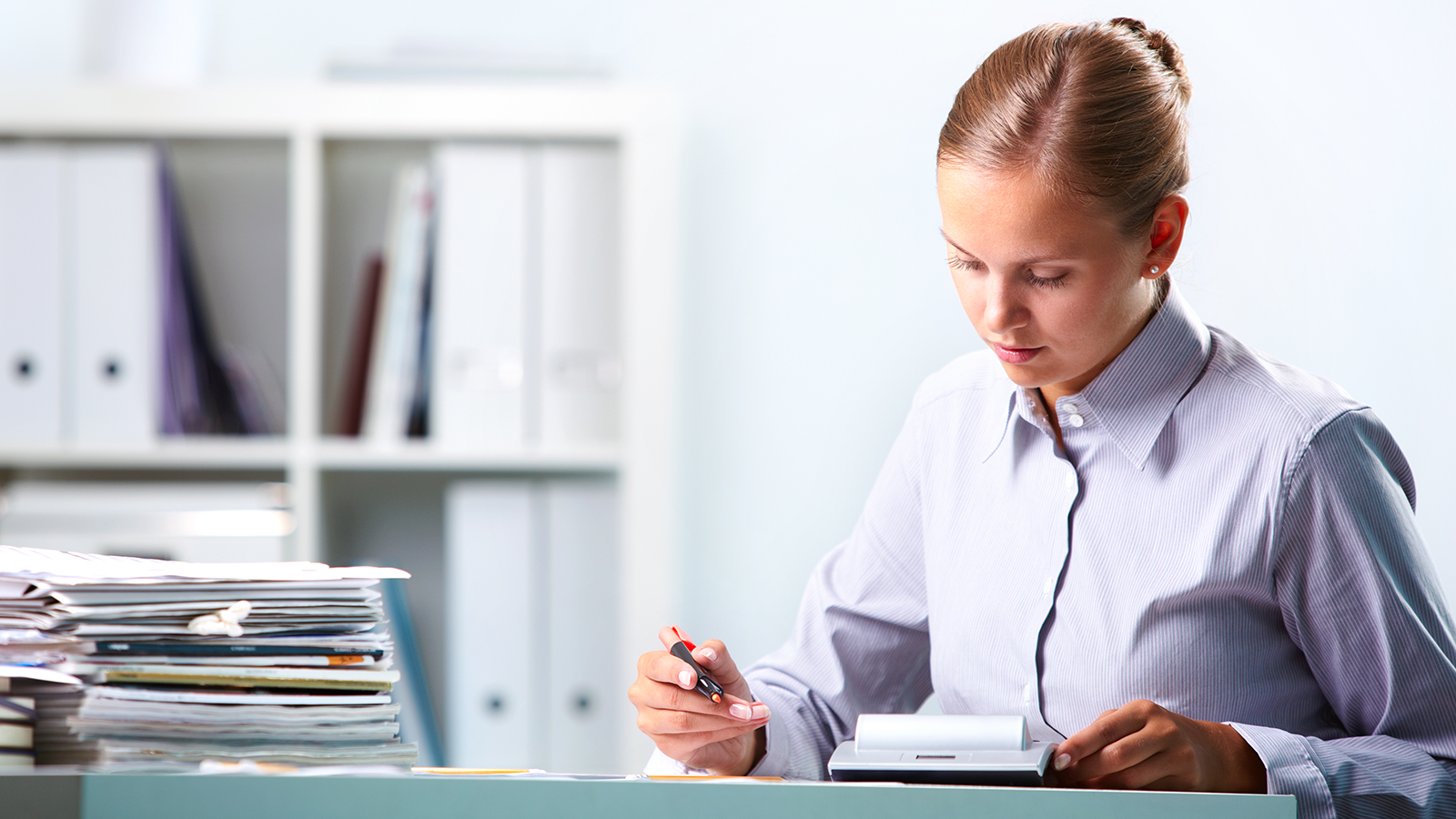 A focused school administrator in a school office working with documents at her desk, surrounded by stacks of paper and binders. The image highlights the need for digital document management to streamline record-keeping and reduce paperwork.