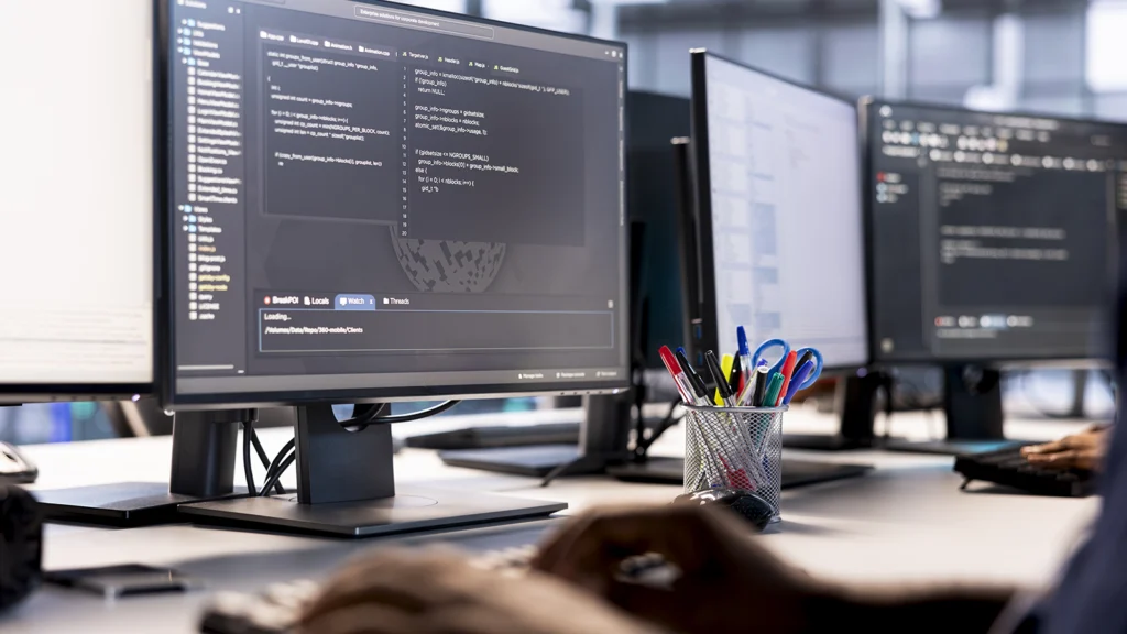 view of various computer screens on a desk. The frontmost monitor is in focus and shows code. Out of focus hands type on a keyboard synced to this computer