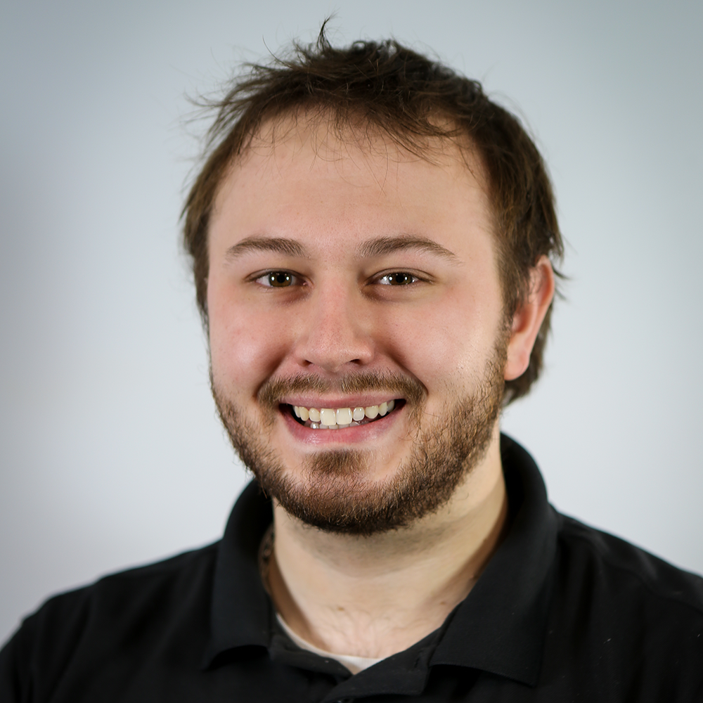 Smiling man with short brown hair and a beard, wearing a black polo shirt against a light gray background.