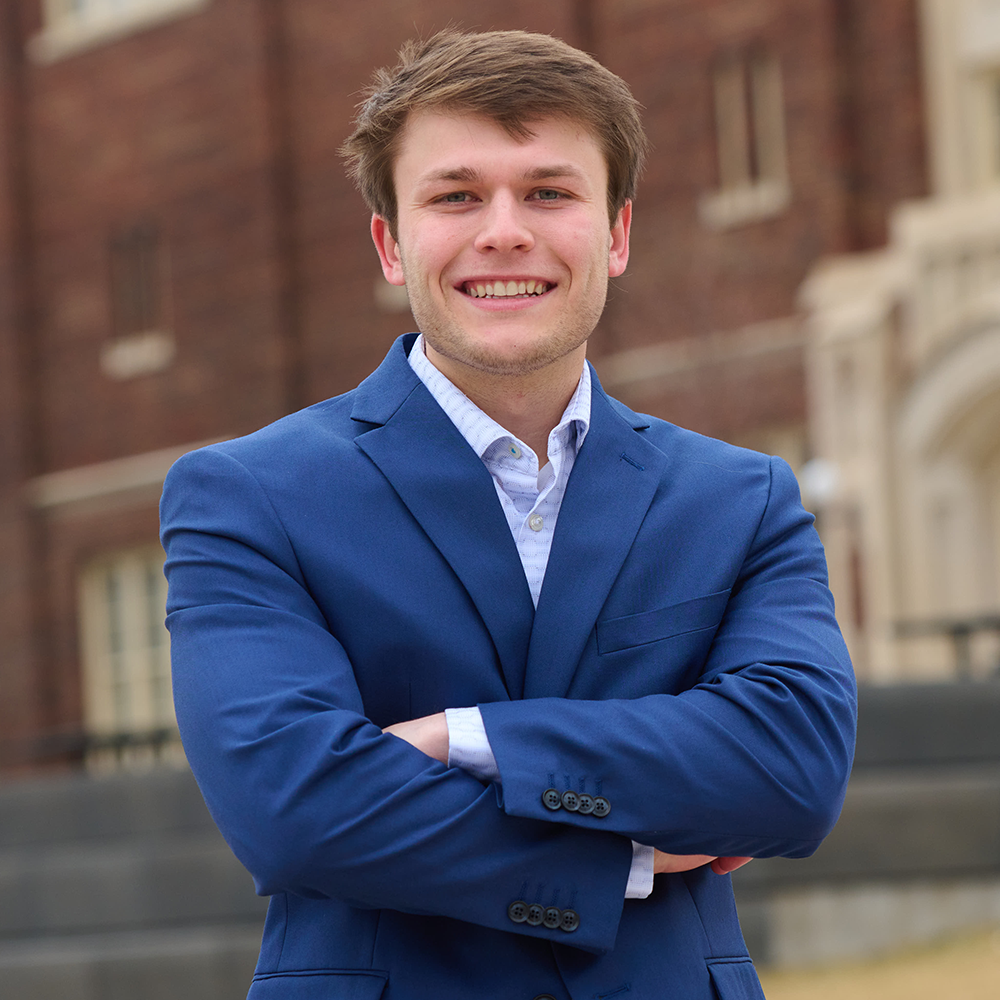 Young man in a blue suit smiling with arms crossed outdoors in front of a brick building.