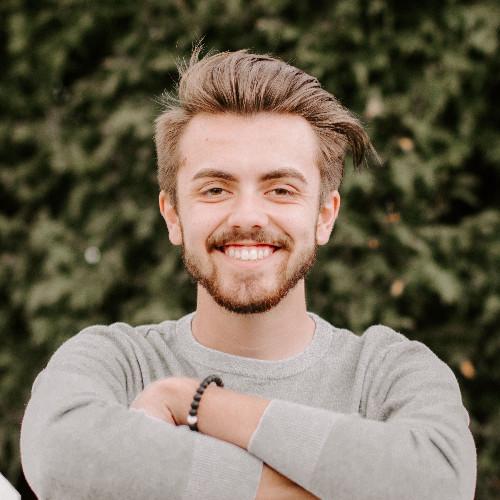 Smiling young man with arms crossed outdoors, wearing a light gray sweater, with greenery in the background.