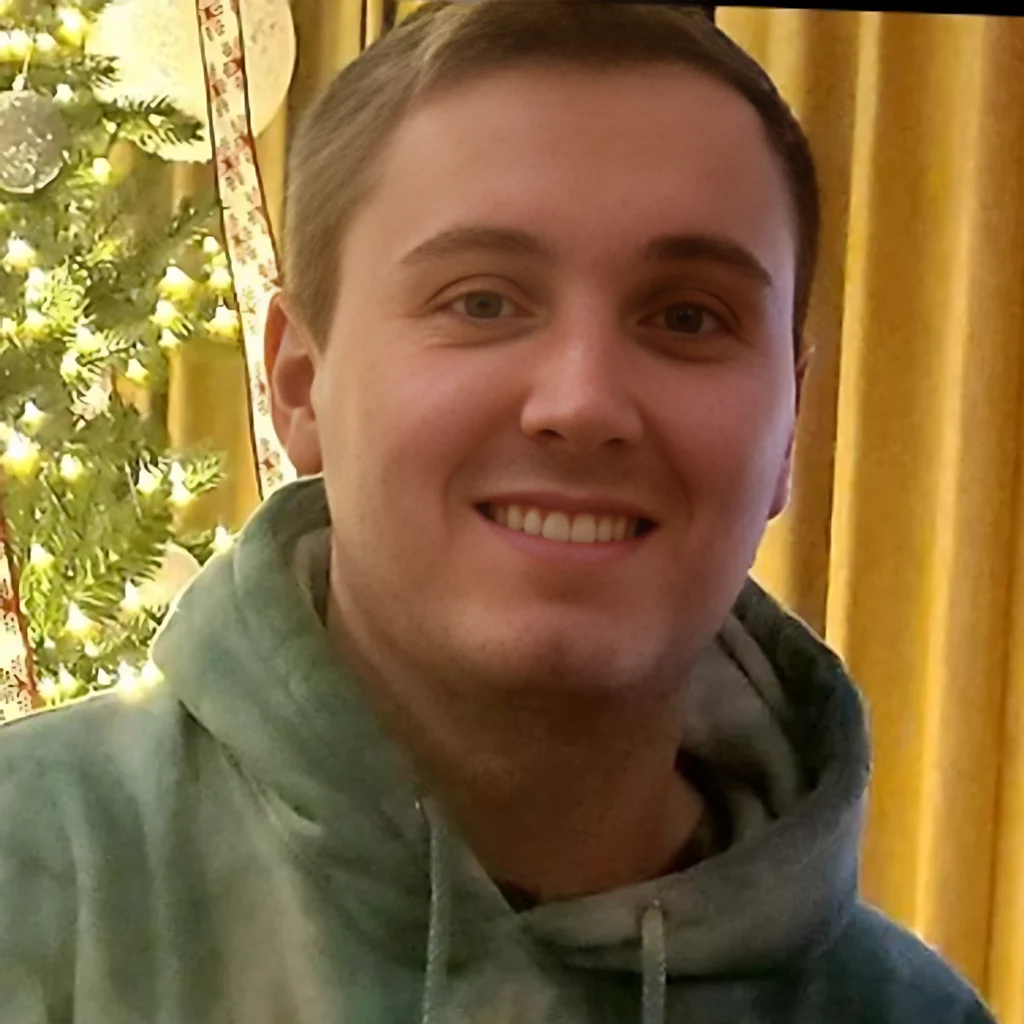 Close-up of a smiling person wearing a hoodie, with warm yellow curtains and greenery in the background (selfie).