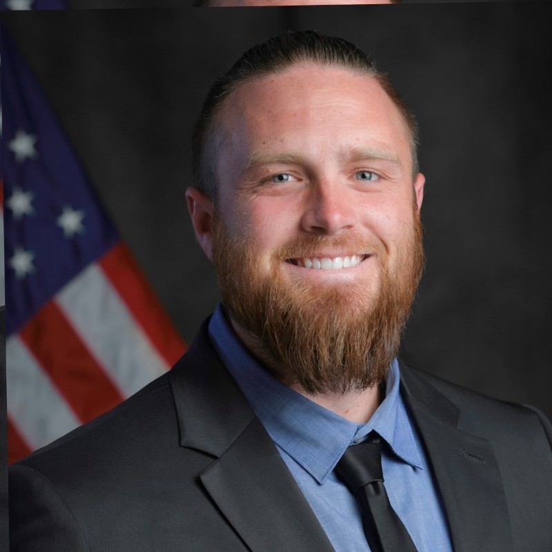 Professional portrait of a smiling man with a full beard in a dark suit and blue shirt, with an American flag in the background.