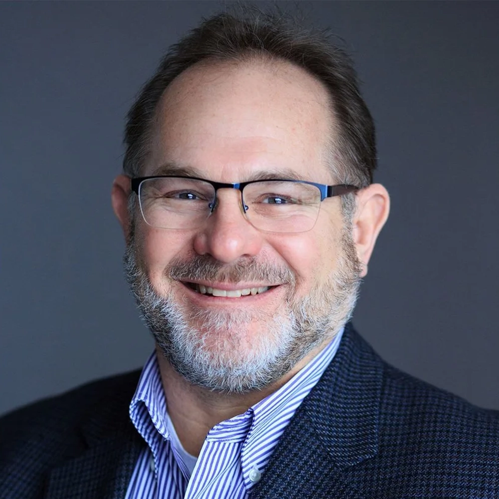Smiling man with glasses and a gray beard in a navy blazer and striped shirt, neutral gray background (professional headshot).