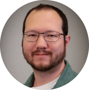 Portrait of a man with short brown hair, glasses, and a beard, wearing a green shirt and white undershirt, smiling slightly