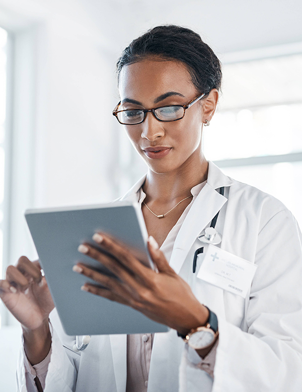 Female doctor in a white coat and glasses reads a tablet in a clinical setting, wearing a stethoscope and name badge.