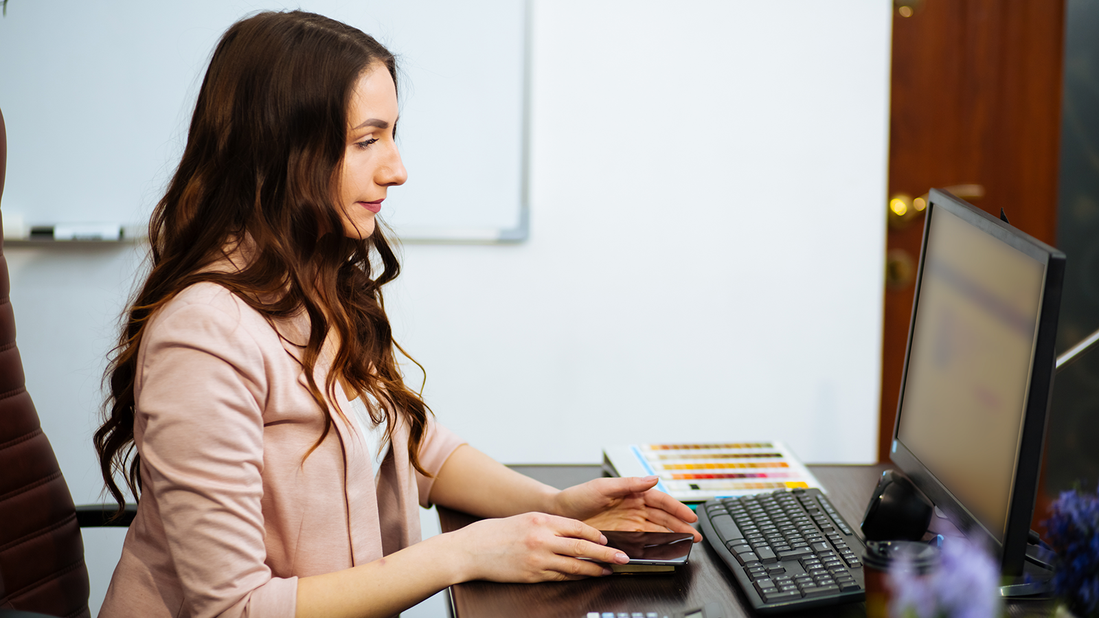 Woman in a pink blazer sits at a desk, using a smartphone beside a computer monitor and keyboard.