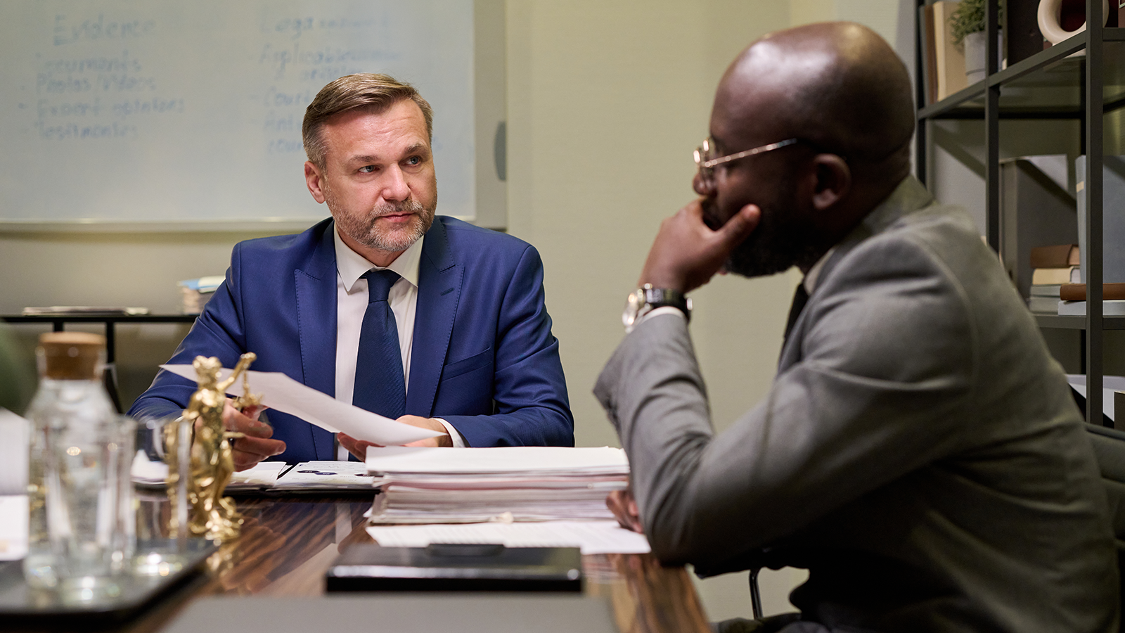 Two businessmen sit across from each other at a desk in a modern office, reviewing documents during a serious meeting.