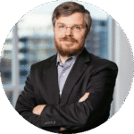 Professional headshot of a bearded man in a dark suit with folded arms, in an office backdrop.