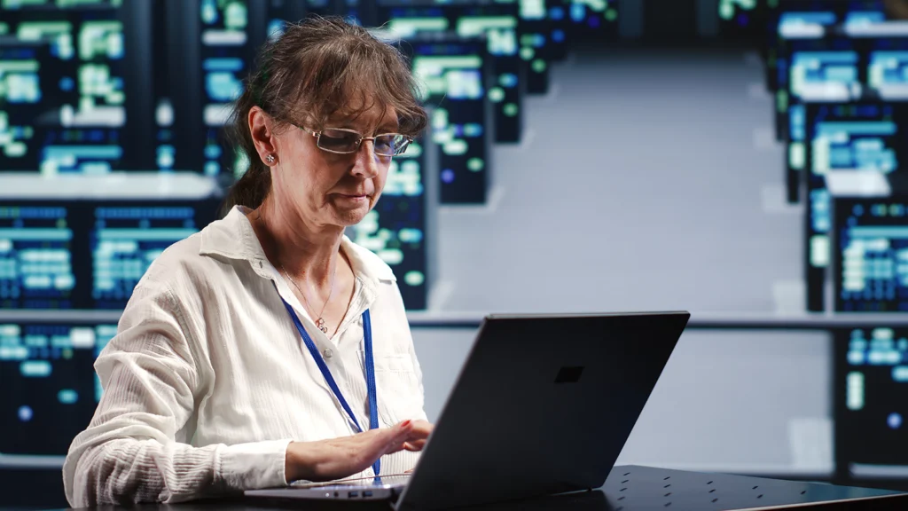 Woman in glasses typing on a laptop in a server room with blue-lit racks at a data center setting.