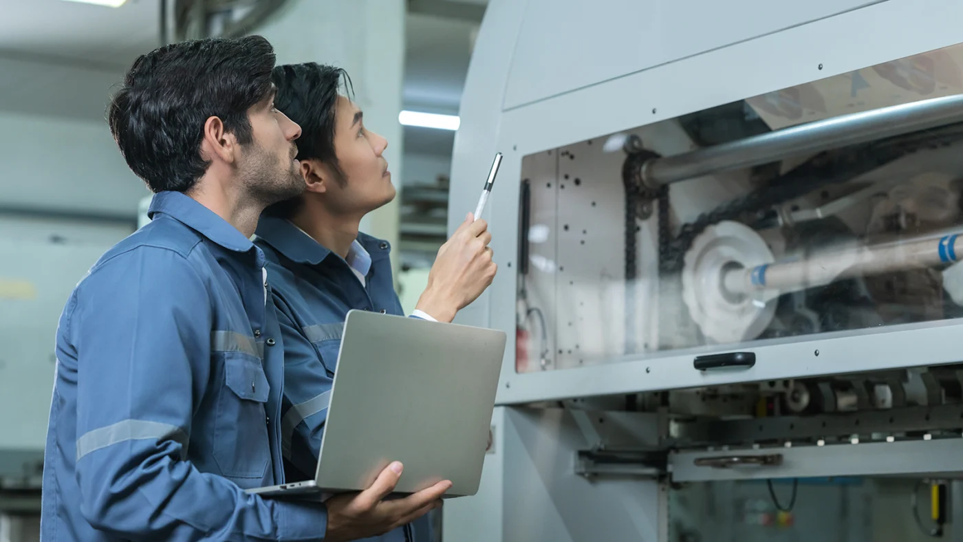 Two technicians in blue uniforms inspect a large industrial machine, one holding a laptop and the other using a pen to examine the equipment nearby in a factory setting.