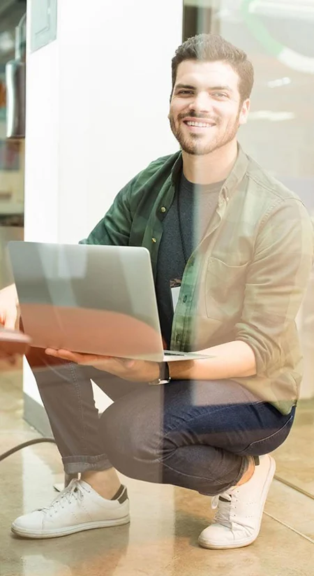 Smiling man crouching on the floor while using a silver laptop outdoors or in a glass-walled space.