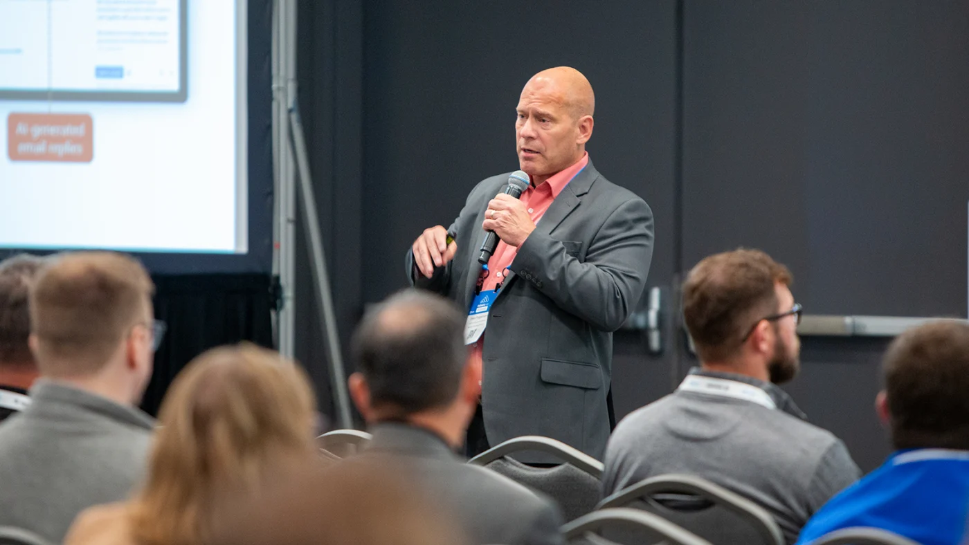 A speaker presenting at the Microsoft Power Platform Envisioning Workshop. The presenter, wearing a suit and holding a microphone, addresses an audience seated in rows while a screen with slides is partially visible in the background.