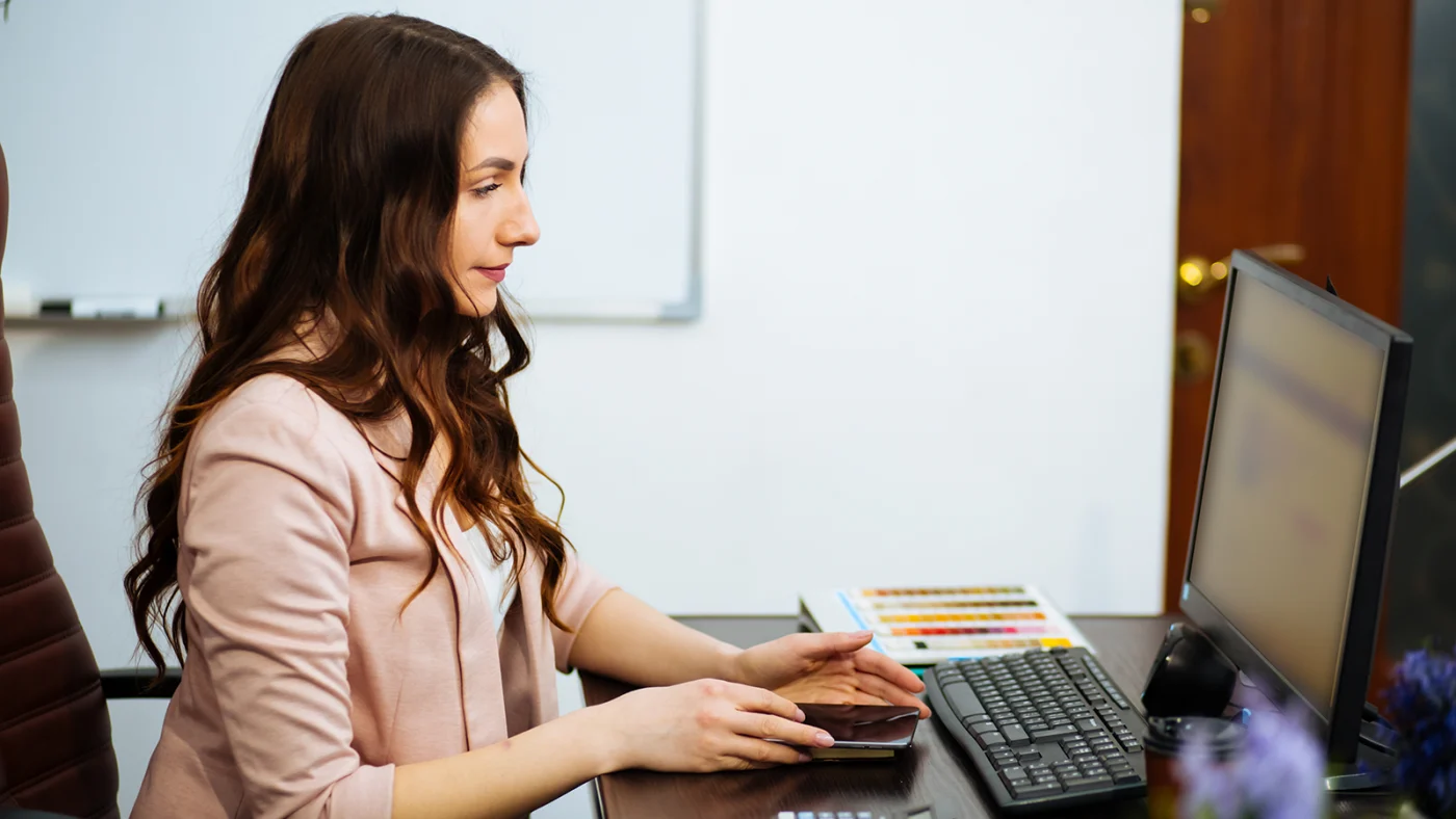 Woman in a pink blazer sits at a desk, using a smartphone beside a computer monitor and keyboard.