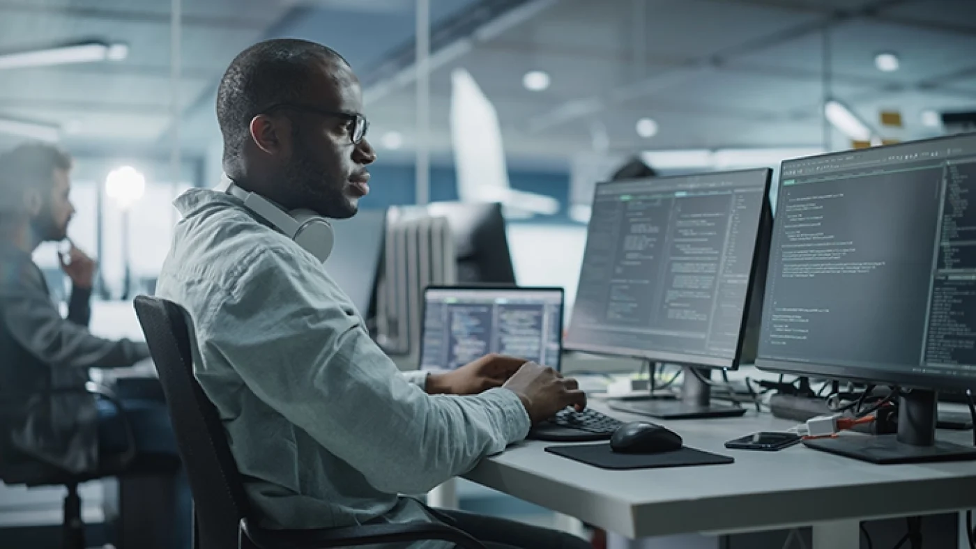 A cybersecurity professional conducts a penetration test on multiple monitors, analyzing code and system vulnerabilities. Another individual is visible in the background working on a similar setup, representing a collaborative environment for vulnerability scanning and pen testing.
